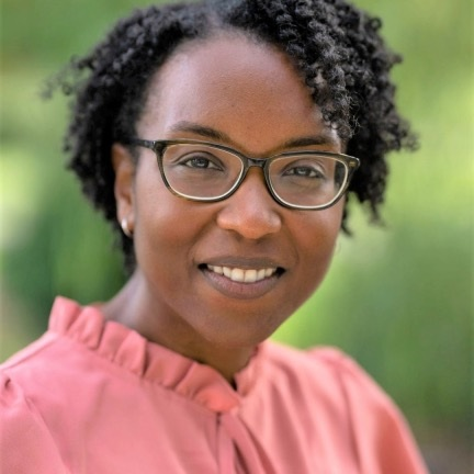 Woman with short curly hair and glasses smiling outdoors in a pink blouse.